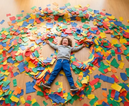 Niño de educación infantil jugando a hacer un ángel en el suelo cubierto de trozos de papel de colores.